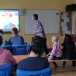 Uczniowie siedzą w ławkach i oglądają prezentację omawianą przez nauczyciela.