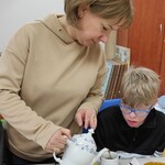 Nauczycielka nalewa z dzbanka do filiżanki herbatę uczniowi siedzącemu przy stole.