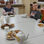 Uczniowie siedzą przy stole na którym stoją talerze z ciastkami, dzbanki z herbatą, filiżanki i talerzyki,