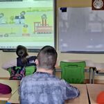 Uczniowie siedzą w ławkach i oglądają prezentację na tablicy multimedialnej.