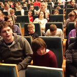 Uczniowie i nauczyciele siedzą na krzesłach kinowych.