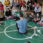 Nauczyciel siedzi na podłodze. Wokół niego siedzą na podłodze uczniowie połączeni nitką trzymaną w dłoniach.
