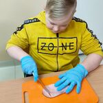 Uczeń siedzi przy ławce. Na ławce znajduje się deska do krojenia, na desce leży wędlina. Uczeń kroi wędlinę.