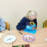 Trzech uczniów siedzi przy swojej ławce. Uczeń po lewej stronie trze ser, dziewczyna w środku trze ser, uczeń z prawej strony kroi paprykę.