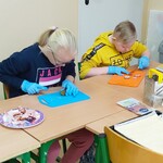 Uczniowie (trzech chłopców i jedna dziewczynka) siedzą przy ławkach. Na ławkach znajdują się deski do krojenia. Uczniowie kroją składniki na pizzę: wędlinę, warzywa.