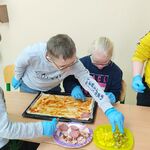 Uczniowie stoją przy ławce. Na ławce znajduje się blacha na której ułożone jest ciasto na pizzę. Uczniowie wspólnie układają produkty na pizzy.