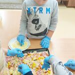 Uczeń stoi przy ławce.  Na ławce znajduje się blacha na której ułożone jest ciasto na pizzę. Uczeń posypuje pizzę serem.