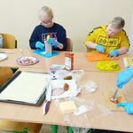 Uczniowie siedzą przy stolikach i kroją produkty na pizzę, trą ser.
