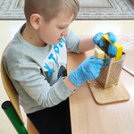 Uczeń siedzi przy ławce. Na ławce stoi tarka. Uczeń trze ser.