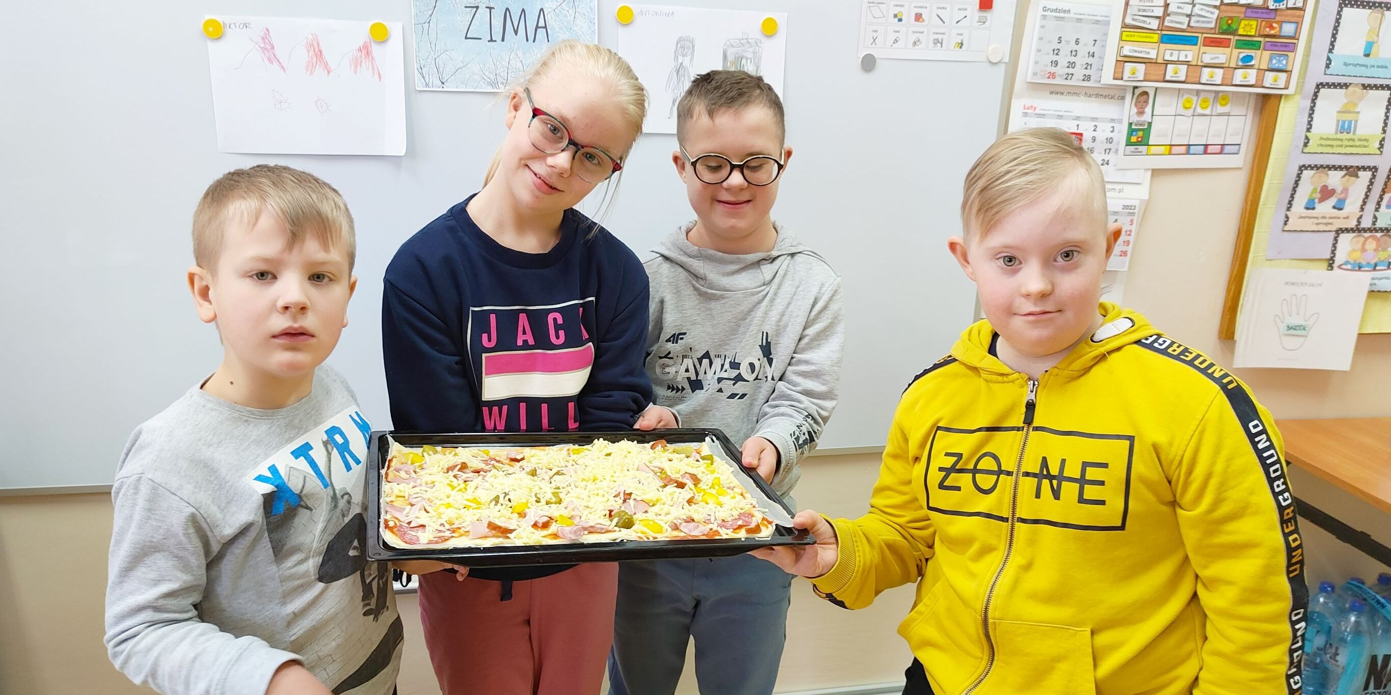 Uczniowie stoją pod tablicą. Wspólnie trzymają przygotowaną przez siebie pizzę.