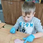 Uczeń siedzi przy ławce. Uczeń siedzi przy ławce. Na ławce znajduje się deska do krojenia, na desce leży wędlina. Uczeń kroi wędlinę.