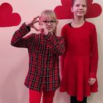 Uczennice w czerwonych ubraniach na tle napisu Walentynki. Niższa dziewczynka pokazuje serduszko z ułożonych dłoni.