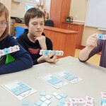 Troje uczniów siedzi przy stole i trzyma w rękach klocki z działaniami matematycznymi,