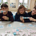 Troje uczniów siedzi przy stole i  trzyma w rękach klocki z działaniami matematycznymi.