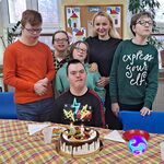 Jubilat siedzi za stołem, na którym stoi tort urodzinowy. Za nim stoi wychowawczyni i koledzy z klasy.