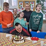Jubilat siedzi za stołem i zdmuchuje świeczkę  na torcie. Za nim stoi wychowawczyni i uczniowie z klasy,