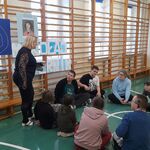 Uczniowie siedzą na podłodze w kółko. Nauczycielka stoi na tle dekoracji zawieszonej na drabinkach.