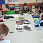 Uczniowie siedzą przy stole i rozwiązują tematyczne karty pracy, Na stole leżą czasopisma i stoi globus.
