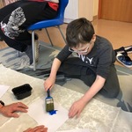 Uczeń siedzi po turecku na podłodze i maluje dużym pędzlem kartkę niebieską farbą.