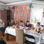 Grupa osób siedzi wokół stołu w udekorowanym pokoju.Na stole znajdują się talerze ze słodkościami urodzinowymi.