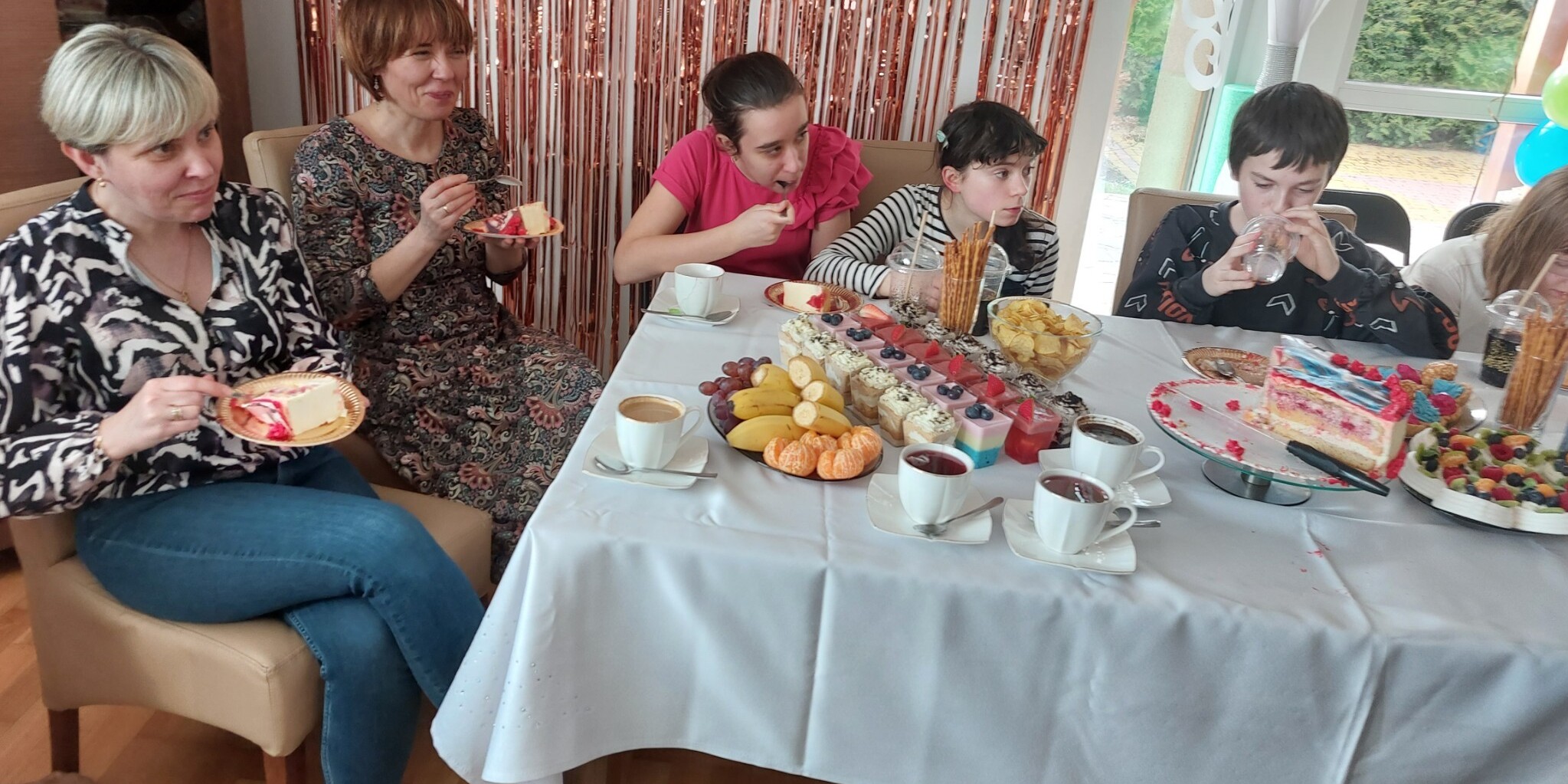Dwie panie i uczniowie siedzą wokół urodzinowego stołu.