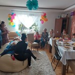 Grupa osób w pokoju siedzi przy stole. Na środku znajduje się duża pufa na której leży jubilat. Obok stoją dwie panie a jedna siedzi na pufie