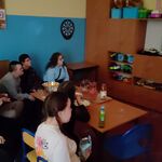 Uczestnicy seansu filmowego siedzą na krzesłach i kanapie i oglądają film. Przed uczniami stoi stół, a na nim znajdują się przekąski (popcorn, ciastka, chrupki) oraz picie.
