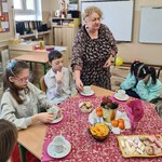 Uczniowie siedzą wokół stołu, Nauczycielka stawia filiżankę na stole przed uczniem.