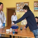 Pani w białym fartuchu stoi przy stole, na którym stoją pojemniki do eksperymentu. Uczeń wlewa barwnik do pojemnika z niebieską wodą.