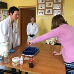 Pani w białym fartuchu stoi przy stole, na którym stoją pojemniki do eksperymentu. Uczennica wsypuje brokat do pojemnika z niebieską wodą.