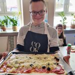 Uczeń trzyma przed sobą dużą blachę z gotową do pieczenia pizzą.