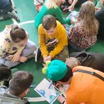 Uczniowie siedzą na podłodze (klasami) i wraz z nauczycielami wykonują zadanie: układają wiosenne puzzle i przyklejają na kartkę.