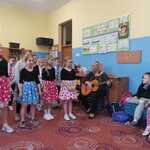 Grupa dziewczynek w spódnicach w kropki stoi. Obok na krześle pani gra na gitarze.