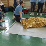 Pani prowadząca zajęcia przykrywa poszkodowaną kocem termicznym. Obok siedzą uczniowie.