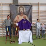 Ksiądz w ornacie.Po bokach stoją uczniowie trzymający się za ręce.