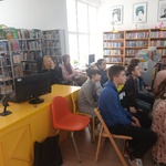 Biblioteczne półki z książkami i uczniowie siedzący na krzesłach.
