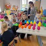 Dzieci siedzą wokół stołu.
Panie patrzą na kolorowe styropianowe jajka na długich patykach, wpięte w kawałek białego styropianu.