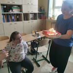 Nauczycielka trzyma tort z płonącymi świeczkami przed uczennicą siedzącą na krześle.