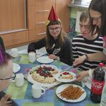 Nauczycielka kroi tort urodzinowy. Wokół stołu siedzą uczniowie w urodzinowych czapeczkach.