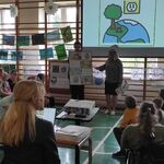 Uczniowie siedzący na ławeczkach patrzą na uczniów i nauczycielkę stojących na tle ekranu ze slajdem.