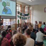 Nauczycielka omawia slajd wyświetlany na ekranie. Uczniowie siedzą wokół na ławeczkach.