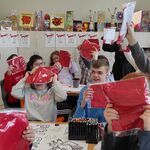 Uczniowie siedzą za stołami. W podniesionych rękach trzymają zapakowane flagi.