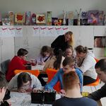 Uczniowie siedzą za stołami w pracowni plastycznej i kolorują mazakami plakaty leżące przed nimi. Z tyłu nauczycielka pochylona nad uczniem.