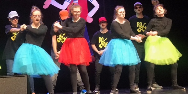 Grupa dzieci w kolorowych strojach tańczy na scenie.