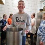 Uczeń trzyma duży garnek do gotowania. W tle grupa dzieci i kucharka.