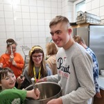Uczeń trzyma duży garnek do gotowania, drugi miesza w garnku chochlą . W tle grupa dzieci i kucharka.
