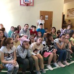 Uczniowie siedzą na ławeczkach, niektórzy maja czapki przedstawiające zwierzęta na głowach.