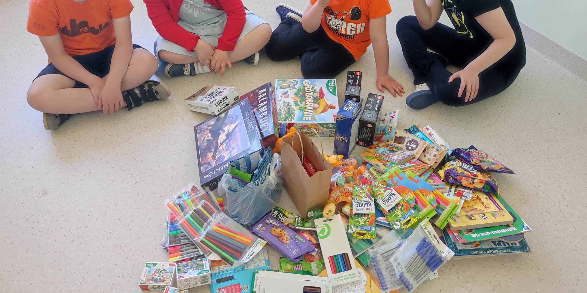 Na podłodze leżą gry, puzzle, słodycze, mazaki, zabawki. Dzieci siedzą wokół zebranych darów.